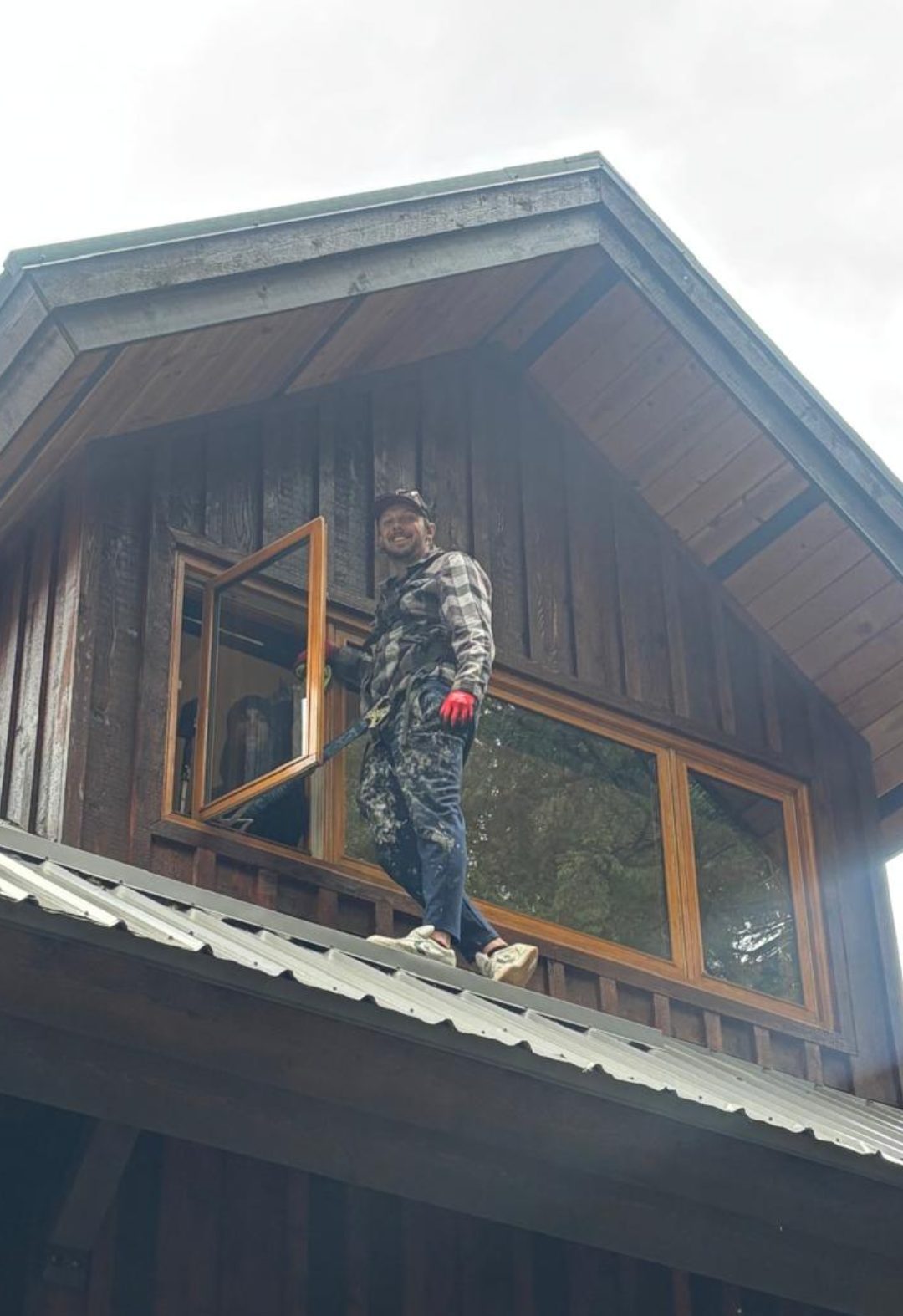 Nigel Purdy smiling on a metal roof in front of a cedar gable he is restaining